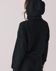 Back view of woman wearing Chaser’s Kiki True Black Zip Up Hoodie with sequin accents with hood up on the shoulders and pockets on a white background
