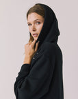 Side view of woman wearing Chaser Aster True Black Hoodie Dress with decorative elements on shoulders with hoodie up on a plain background