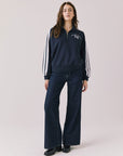 Woman wearing Chaser's navy Atelier Sports 76 Half Zip Long Sleeve with white stripes and dark denim jeans on a light gray background
