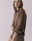 Side view of a woman wearing the Chaser brown All Day Cognac Wash Button Down with matching Sloane Mini Skirt against a plain background