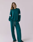 Full body front view of a woman wearing Chaser's Cornell Pullover in Teal and White with striped sleeves and the matching Sandi Joggers on a plain studio background