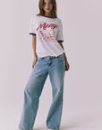 Front view of a woman wearing Chaser's white Desert Mirage Motor Club Tee with red text and graphics and blue jeans on a plain background