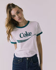 Front view of a woman wearing Chaser's white Coca Cola The Real Thing tee with 'Coke' logo and blue jeans on a plain background