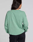 A woman viewed from the back wearing a sage green long-sleeve top and dark jeans against a light gray studio background.