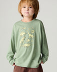 Front view of a boy wearing the green Chaser Dino Action Boys Tee on a neutral background with pants
