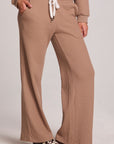Front view of a woman wearing the Chaser Jorgia Camel Pants on a neutral background with shoes and hands in pocket