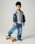 Front view of a boy wearing the Chaser Team Spirit Boys Jacket on a neutral background with jeans and white shirt riding a skateboard