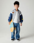 Front view of a boy wearing the Chaser Team Spirit Boys Jacket on a neutral background with jeans and white shirt holding a skateboard under one arm