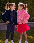 Chaser Kids Avalon Girls Puff-Sleeve Jacket in navy and Camelia Girls Puff-Sleeve Jacket in pink, worn by two girls on grass beside a low wall and greenery