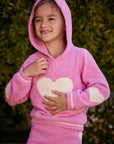 Chaser Kids Holiday Hug Girls Hoodie in pink with white hearts, close-up with hood up outdoors in front of greenery