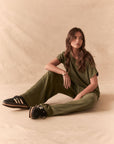 Full front view of women sitting wearing Chaser's Lee Trouser in bronze green with wide-leg silhouette and relaxed drape with matching tee against a beige background