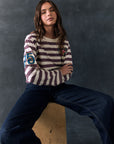 Full body view of model sitting on a wooden block against a dark background wearing The Rolling Stones Rugby Stones Logo Long Sleeve with red and white stripes, cropped fit, tongue logo graphic, and jeans