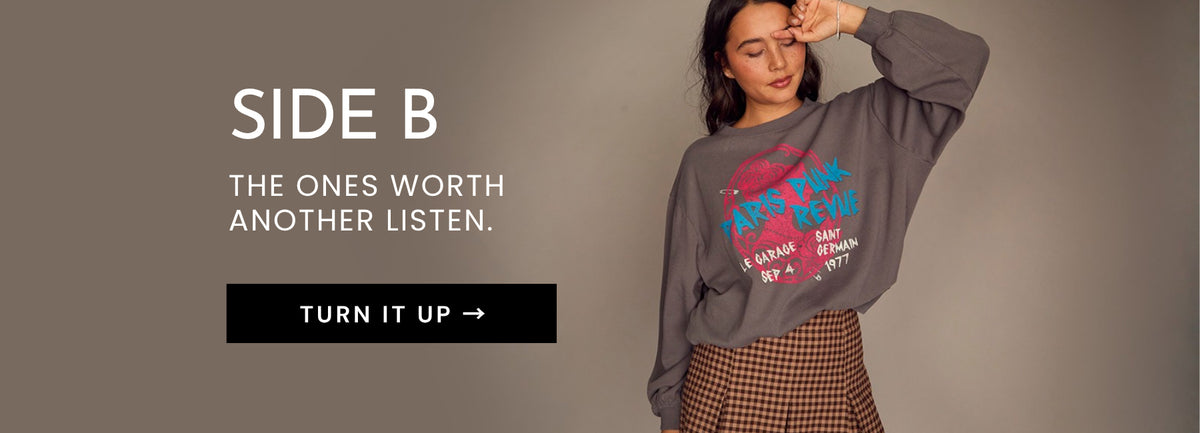 A woman wearing a dark gray graphic sweatshirt and a brown checkered skirt stands against a plain taupe background, with large text on the left reading 'SIDE B' and 'THE ONES WORTH ANOTHER LISTEN.' and a black 'TURN IT UP' button