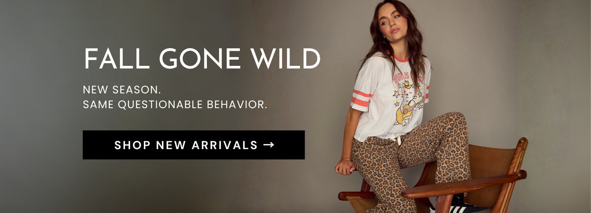 A fashion advertisement featuring a woman sitting on a wooden chair, wearing a white graphic t-shirt with pink stripes and leopard-print pants. The left side of the image has text that reads 'FALL GONE WILD' and 'SHOP NEW ARRIVALS ->'