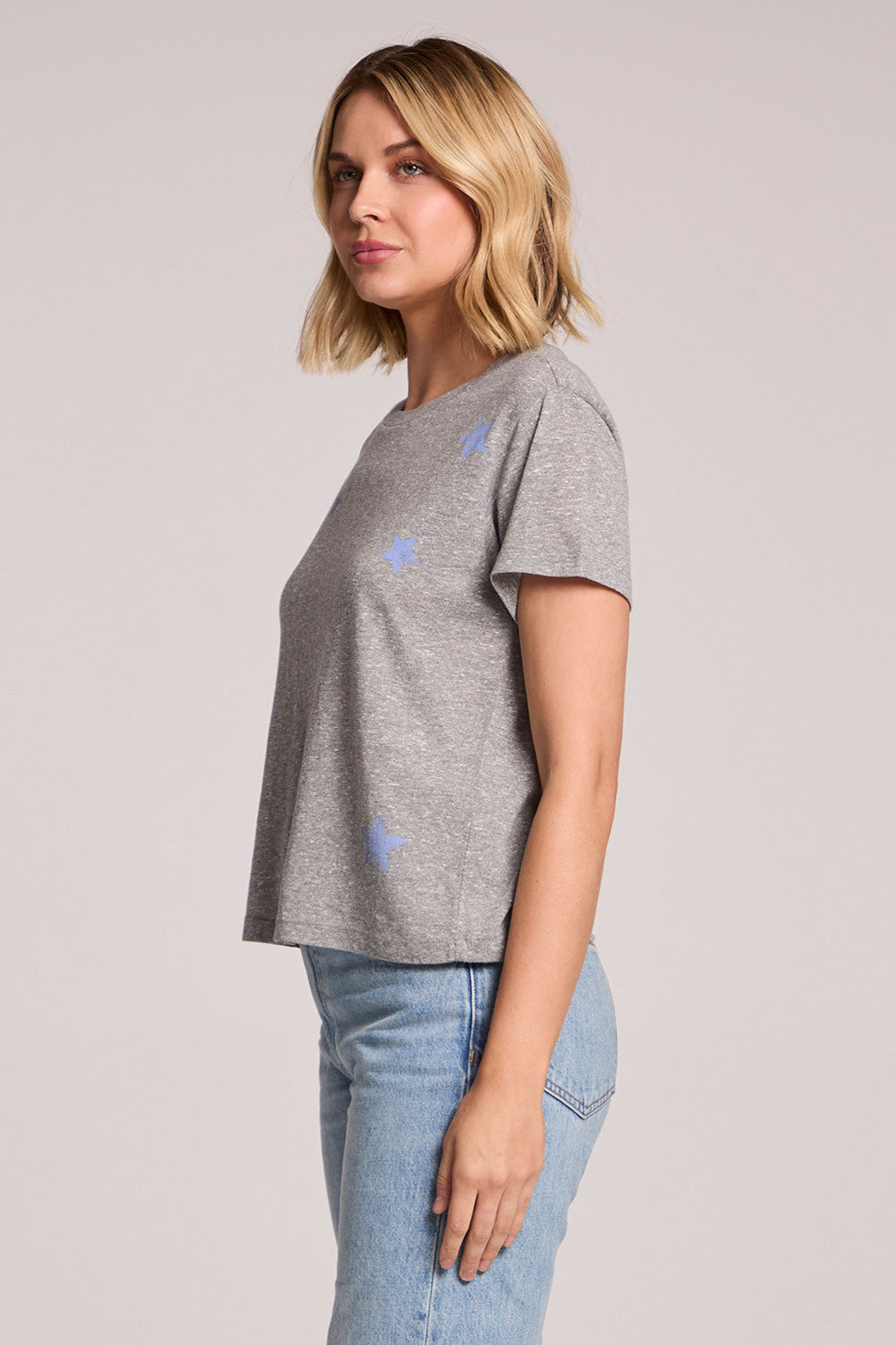 Side view of a woman wearing the Chaser  Allover Stars Tee on a neutral background with her arms at her side