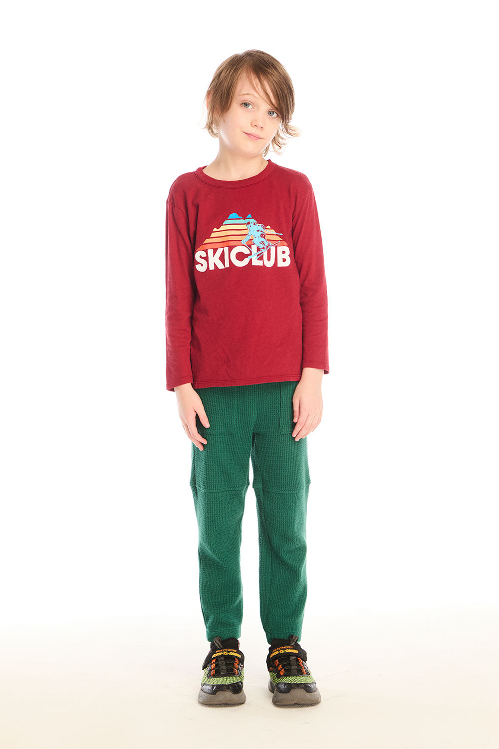 Full body view of boy wearing Chaser’s Ski Club red pullover long sleeve with SKICLUB mountain graphic and green pants on a white background
