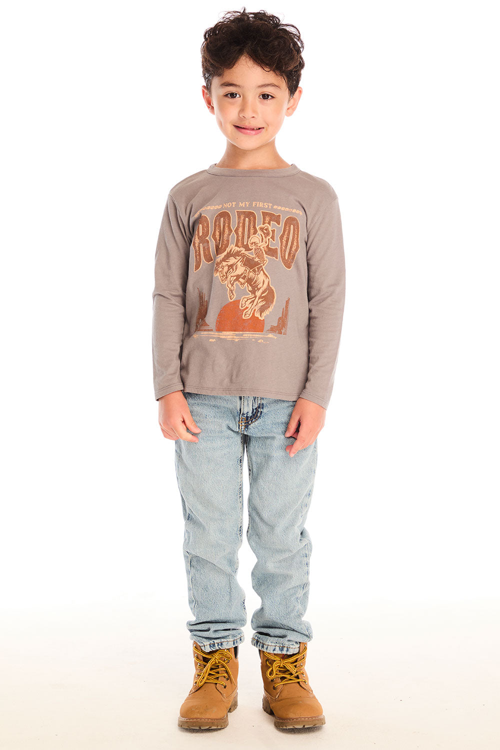 Full body view of boy wearing Chaser’s Hold Your Horses dark grey long sleeve with cowboy graphic, jeans, and boots on a white background