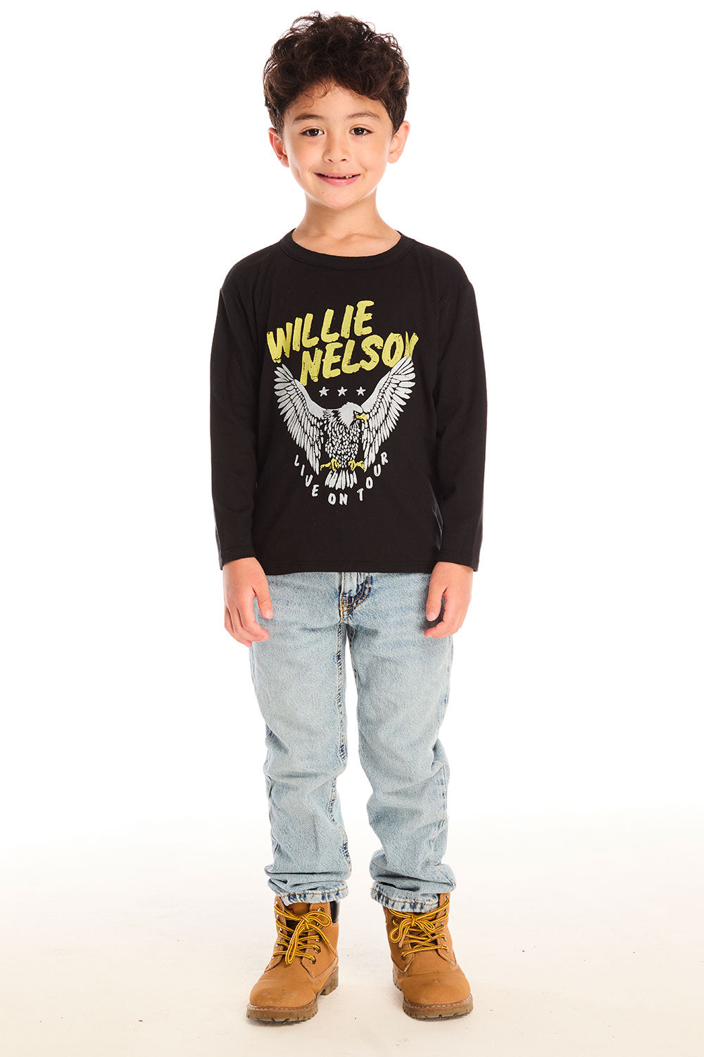 Full body view of boy wearing Chaser’s Willie Nelson Willie 1978 black long sleeve with eagle graphic, light blue jeans, and brown boots on a white background