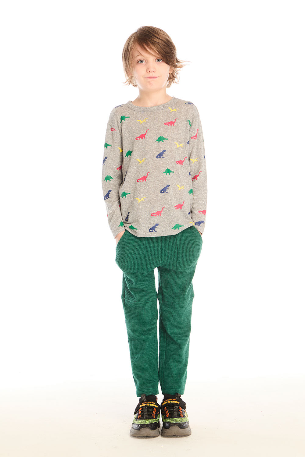  Full body view of boy wearing Chaser’s Multi Dinos grey long sleeve top with colorful dinosaur patterns and green pants on a white background
