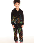 Full body view of boy wearing Chaser’s Paint Splatter black zip up hoodie with colorful splatter design, matching bottoms, and red sneakers on a white background