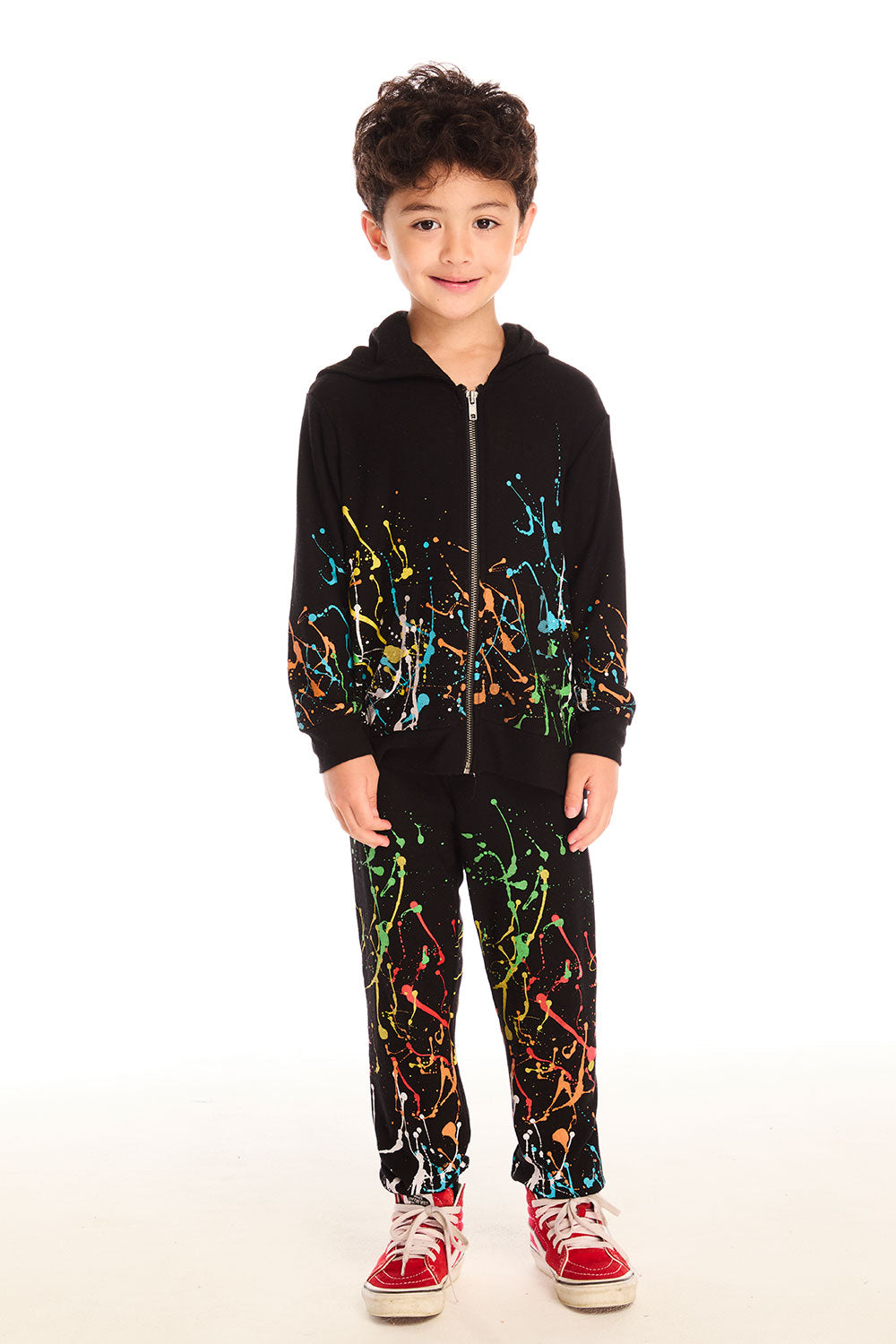 Full body view of boy wearing Chaser’s Paint Splatter black pants with colorful splatter design, matching zip up hoodie, and red sneakers on a white background