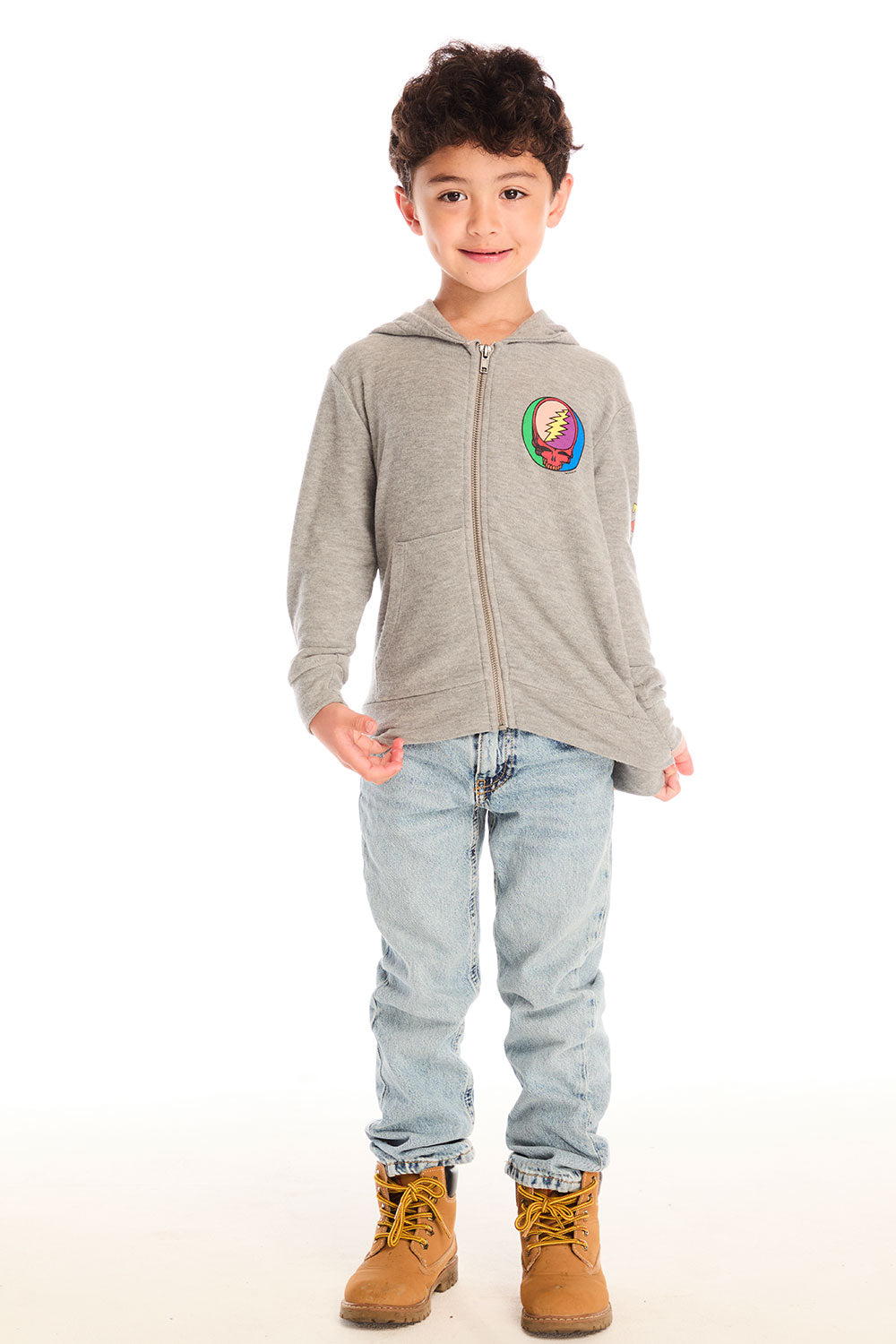 Full body view of boy wearing Chaser’s The Grateful Dead Colorblocked Stealie heather grey zip up hoodie with colorful Steal Your Face logo, light blue jeans, and brown boots on a white background