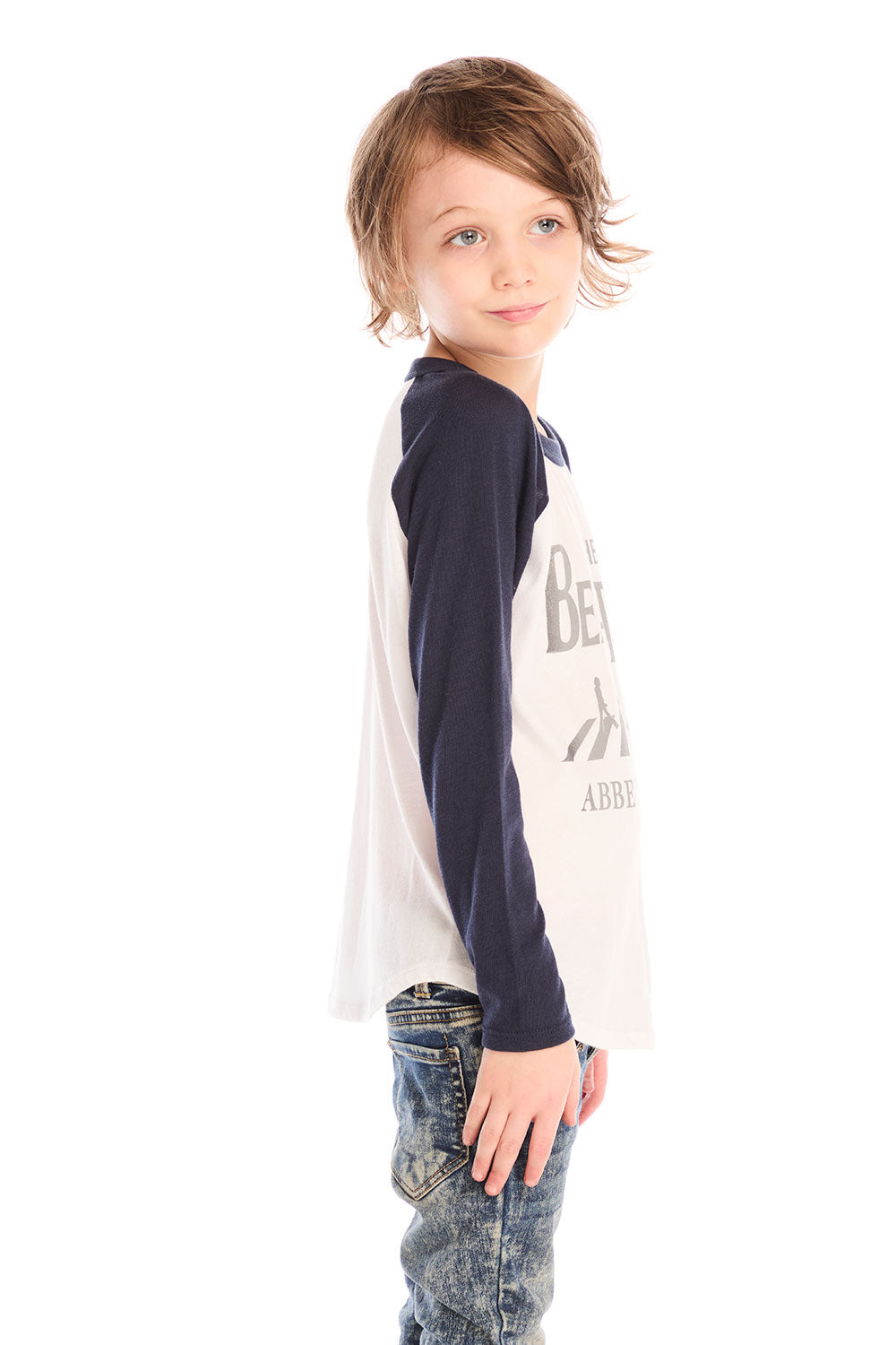 Side view of boy wearing Chaser’s The Beatles Abbey Road football tee in white with navy blue sleeves and jeans on a white background