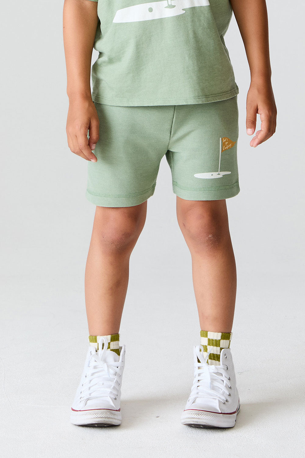 Close up view of boy wearing Chaser Kids Lets go Golfing shorts with white sneakers on a white background