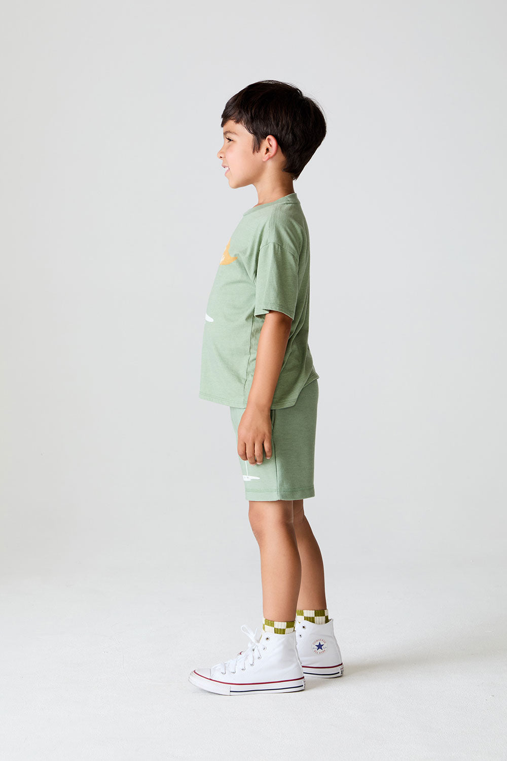Side view of boy wearing Chaser Kids Lets go Golfing shorts and matching green t-shirt with white sneakers on a white background