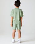 Back view of boy wearing Chaser Kids Lets go Golfing shorts and matching green t-shirt with white sneakers on a white background