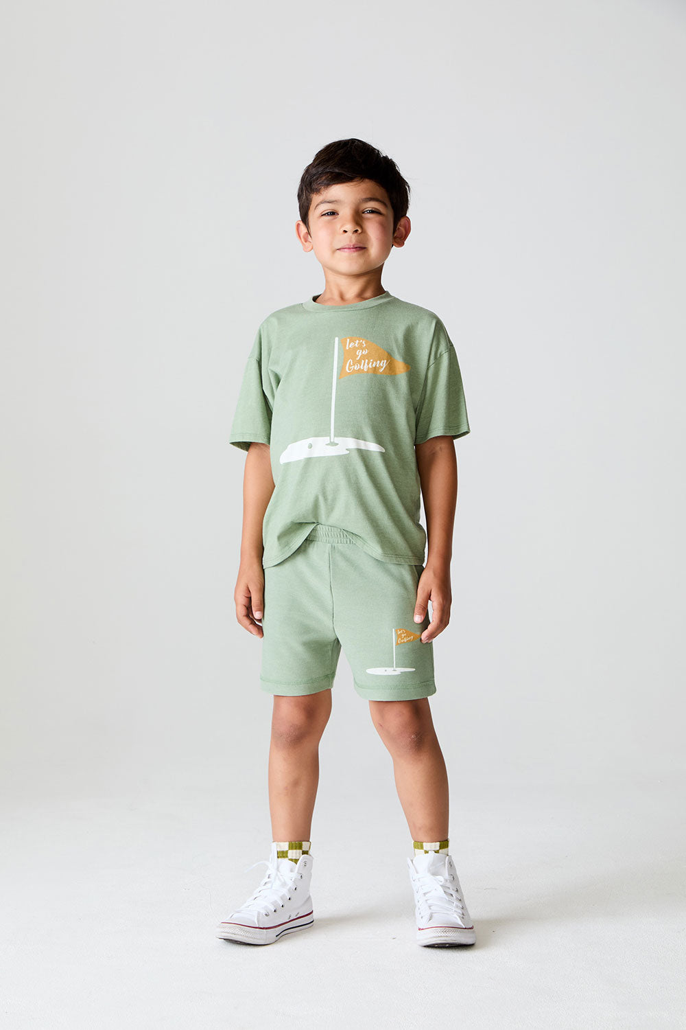 Full view of boy wearing Chaser Kids Lets go Golfing shorts and matching green t-shirt with white sneakers on a white background