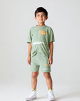 Full view of boy wearing Chaser Kids Lets go Golfing shorts and matching green t-shirt with white sneakers on a white background