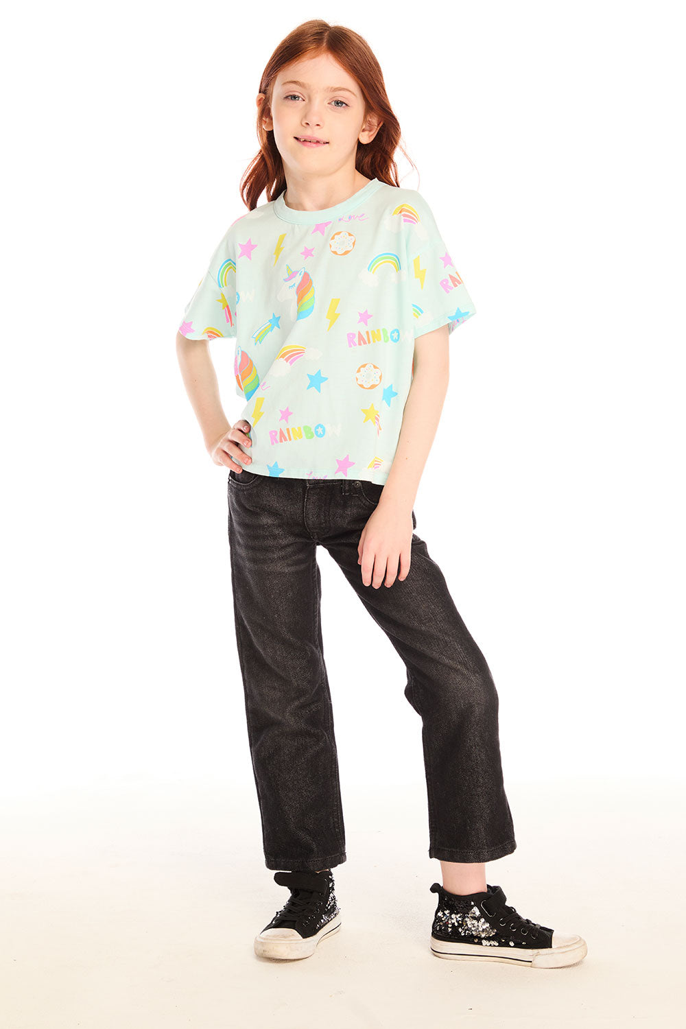 Full body view of girl wearing Chaser’s light green Unicorn Hearts Tee with text and colorful graphics, black jeans, and shiny sneakers against a white background