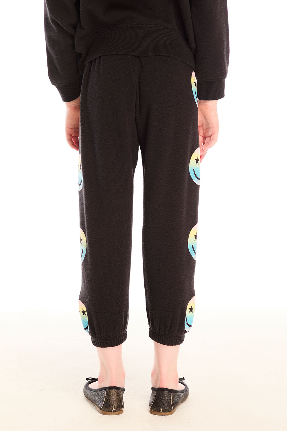 Back view of girl wearing Chaser’s black Pastel Rainbow Smiley Pants with pastel-colored smiley face designs on a white background
