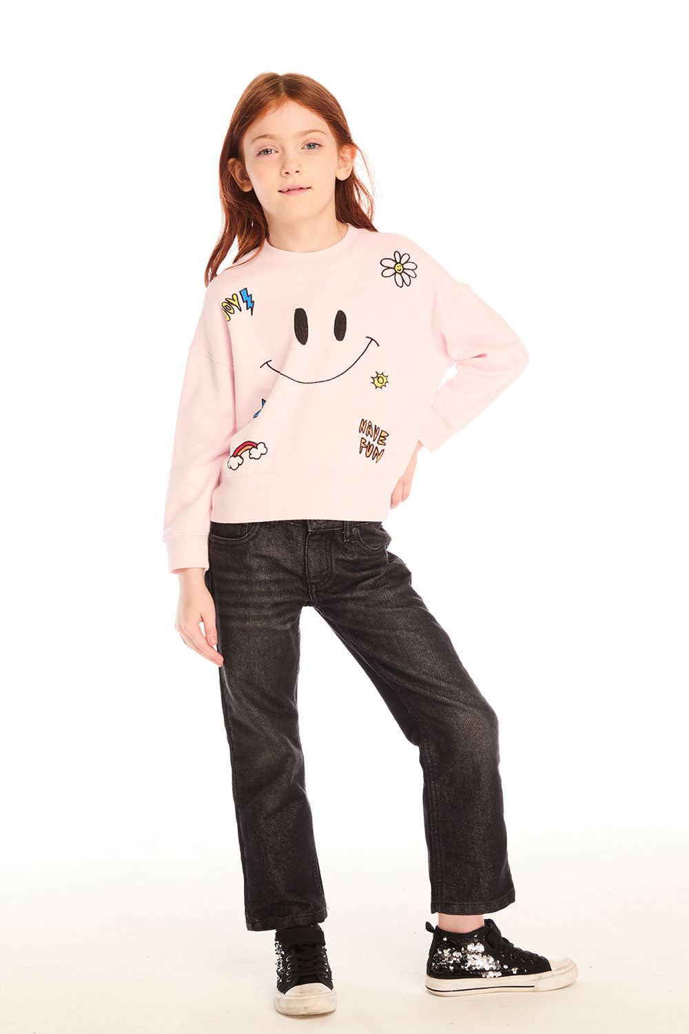 Full body view of young girl wearing Chaser’s pink Be Happy Icons long sleeve pullover with smiley face design and black jeans on a white background