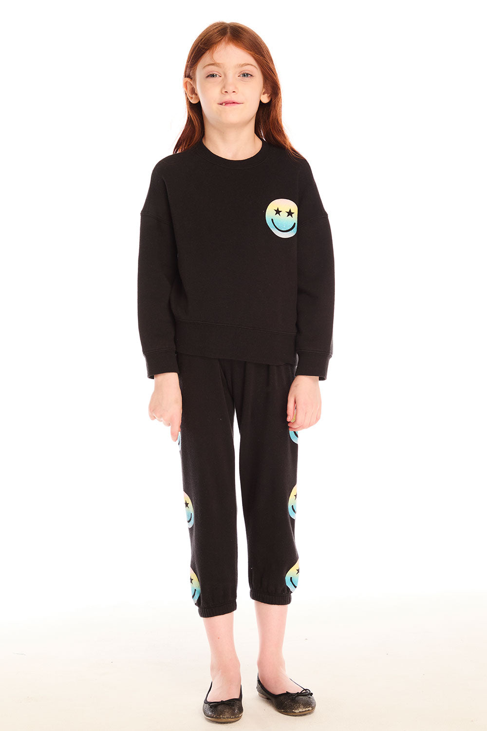 Full body view of girl wearing Chaser’s black Pastel Rainbow Smiley Pants with pastel-colored smiley face designs and matching top on a white background