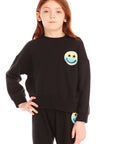 Girl wearing Chaser’s black Pastel Rainbow Smiley Long Sleeve Top with pastel-colored smiley face designs on a white background