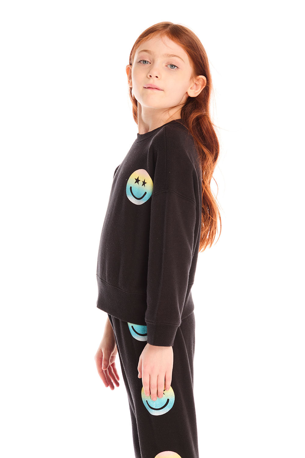 Side view of girl wearing Chaser’s black Pastel Rainbow Smiley Long Sleeve Top with pastel-colored smiley face designs on a white background