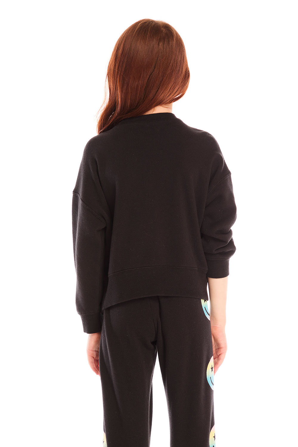 Back view of girl wearing Chaser’s black Pastel Rainbow Smiley Long Sleeve Top with pastel-colored smiley face designs on a white background
