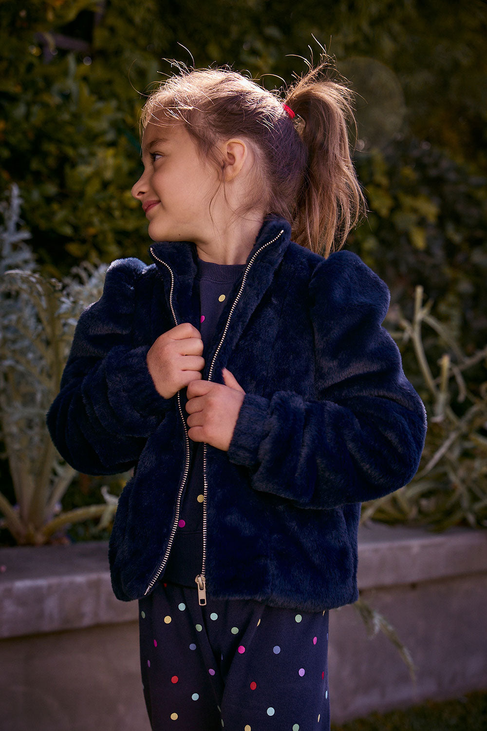 Chaser Kids Avalon Girls Puff-Sleeve Jacket in navy plush fabric, front three-quarter view outdoors beside a low wall and greenery