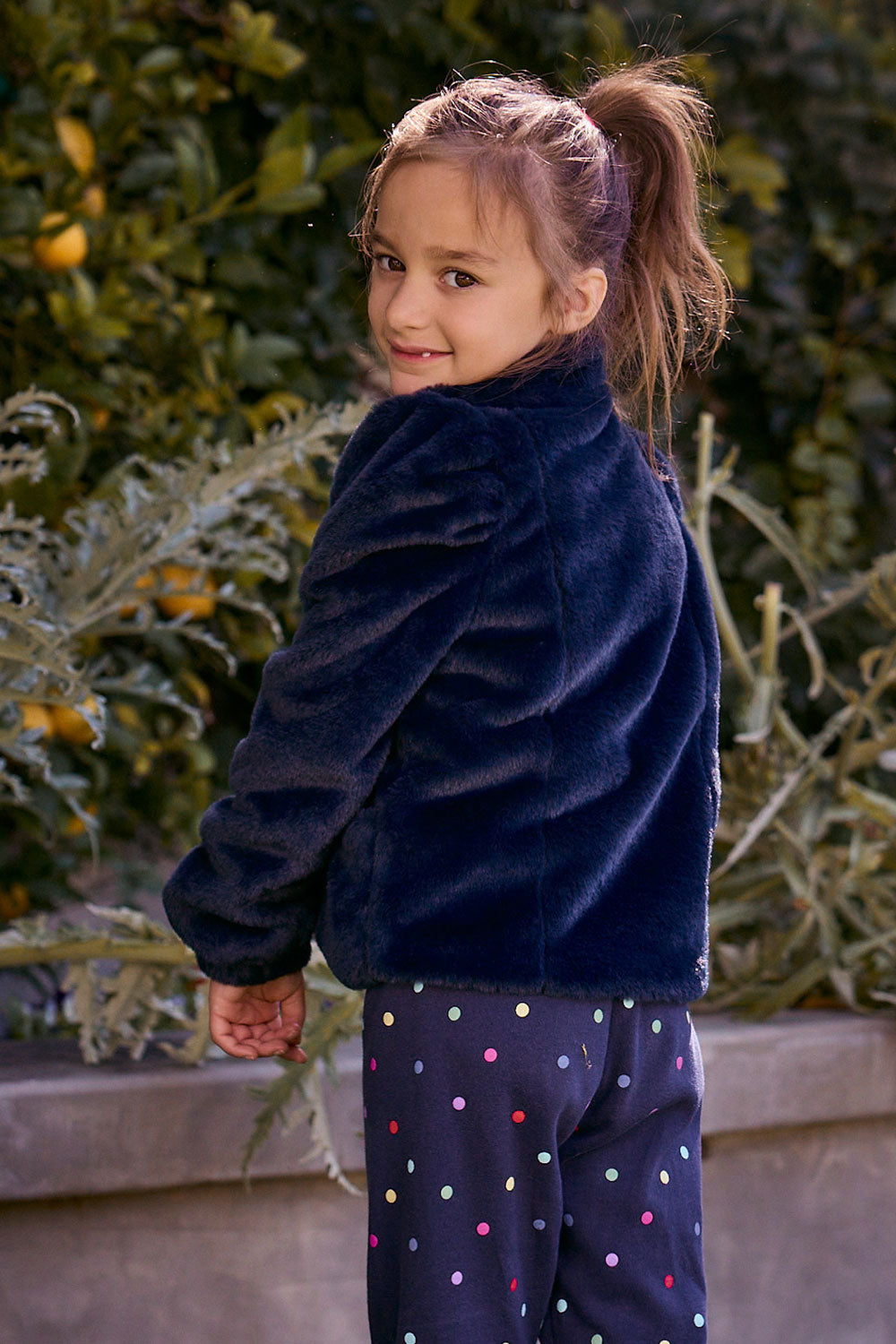 Chaser Kids Avalon Girls Puff-Sleeve Jacket in navy plush fabric, rear three-quarter view outdoors beside a low wall and greenery