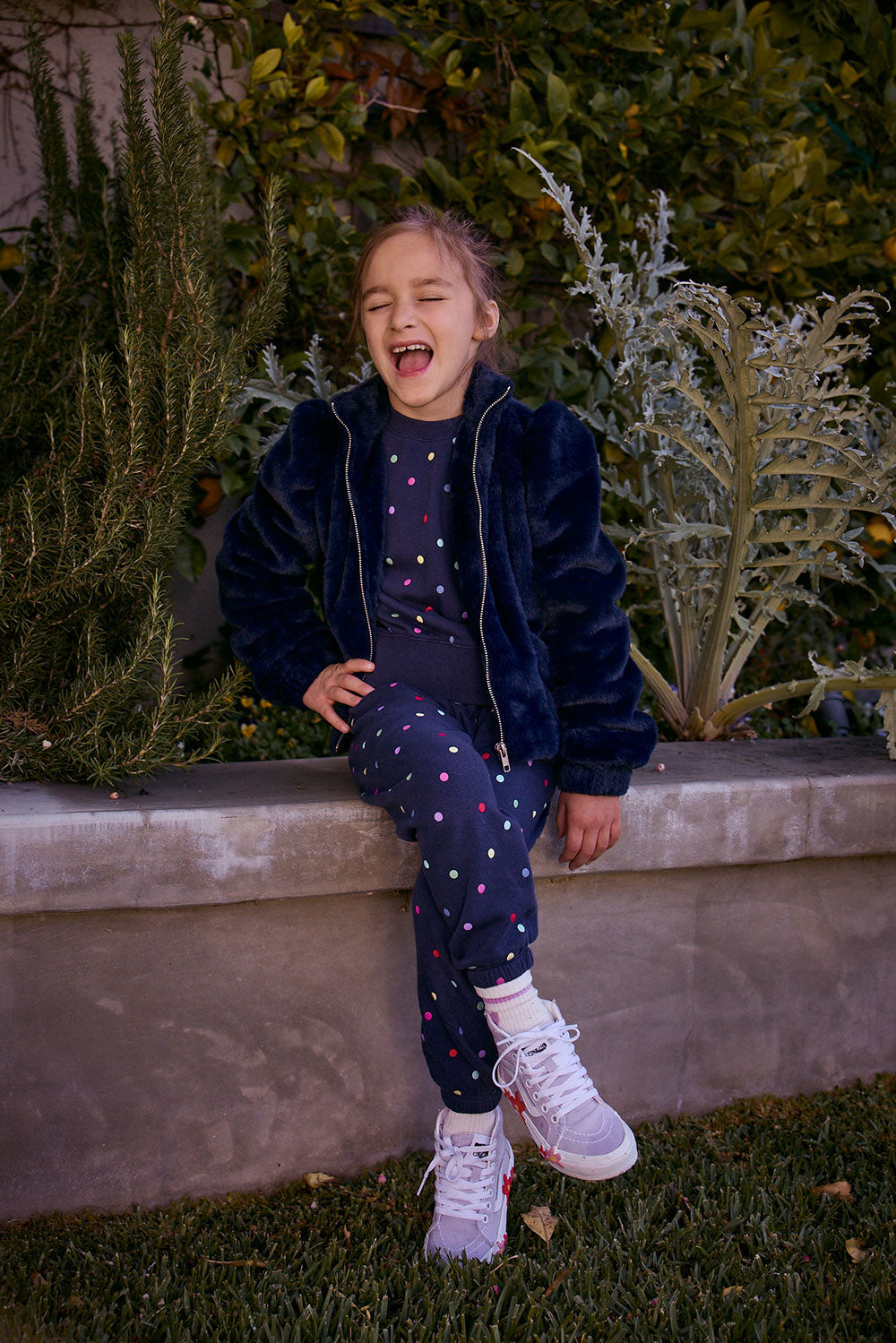Chaser Kids Avalon Girls Puff-Sleeve Jacket in navy, full-body view of a girl seated on a low wall outdoors beside greenery