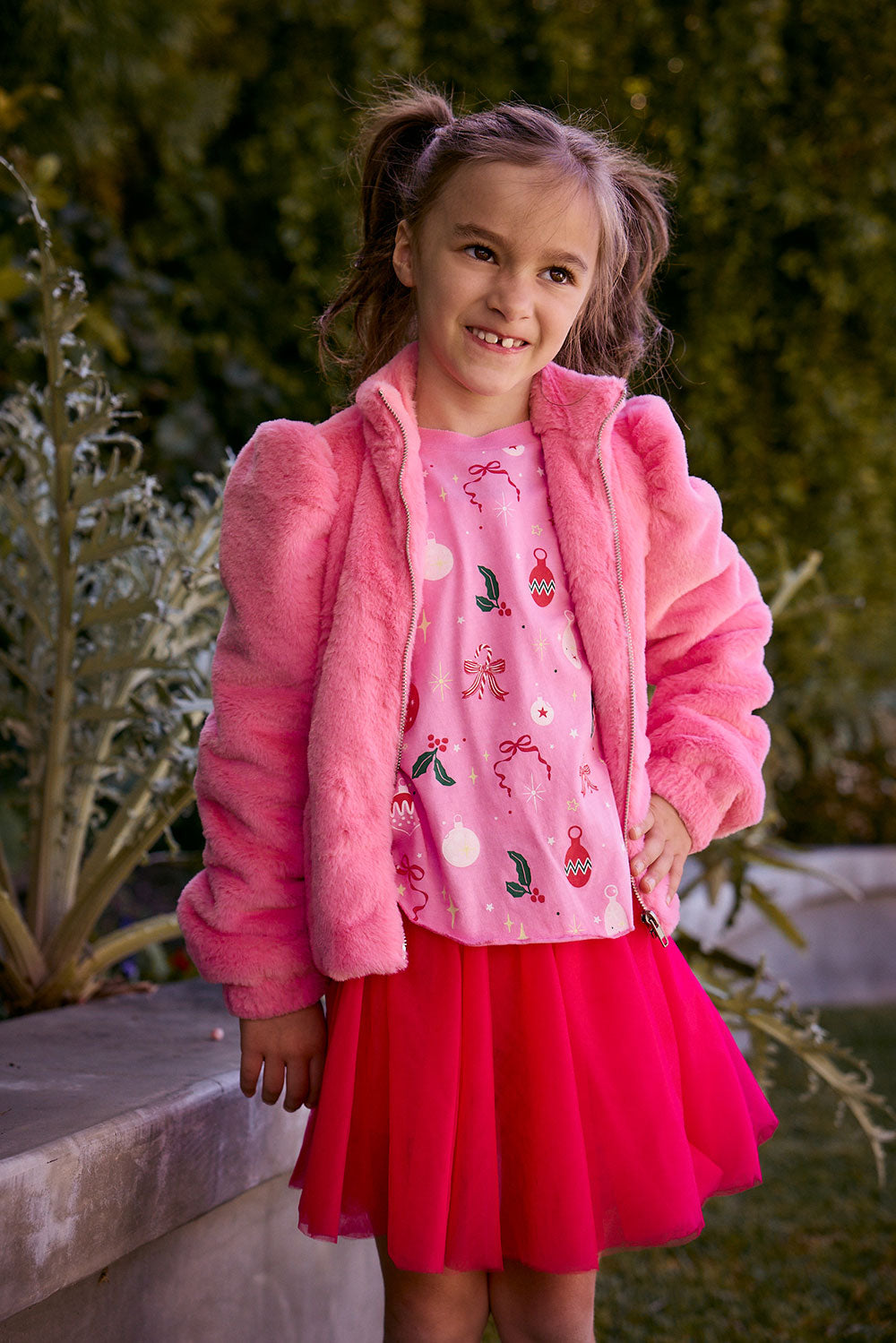 Chaser Kids Camelia Girls Puff-Sleeve Jacket in pink plush fabric, worn open, front view outdoors beside a low wall and greenery