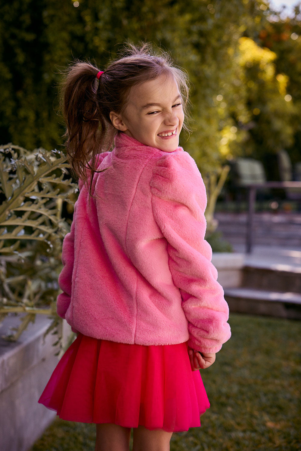 Chaser Kids Camelia Girls Puff-Sleeve Jacket in pink plush fabric, rear three-quarter view outdoors beside a low wall and greenery