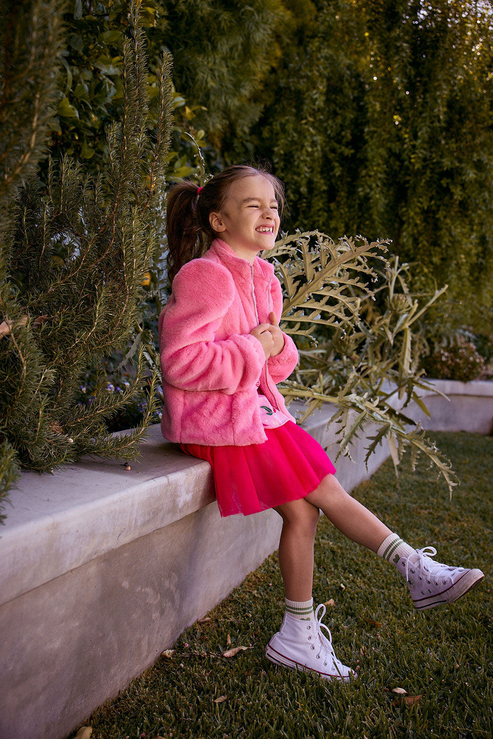 Chaser Kids Camelia Girls Puff-Sleeve Jacket in pink, full-body view of a girl seated on a low wall outdoors beside greenery