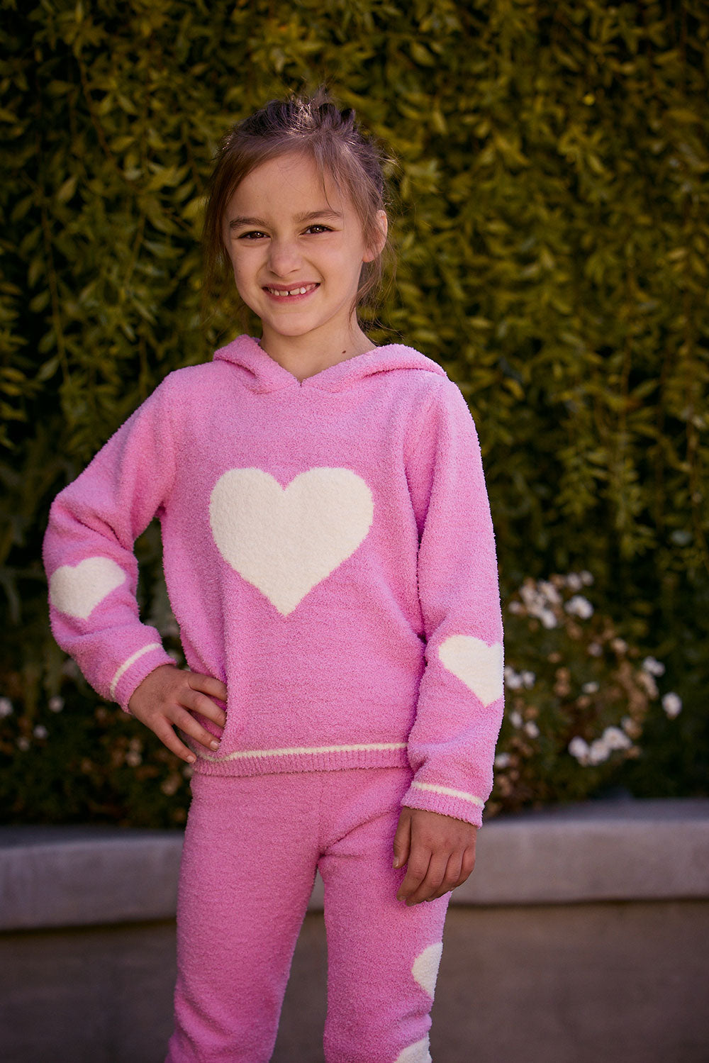 Chaser Kids Holiday Hug Girls Hoodie in pink with white hearts, front view outdoors beside a low wall and greenery