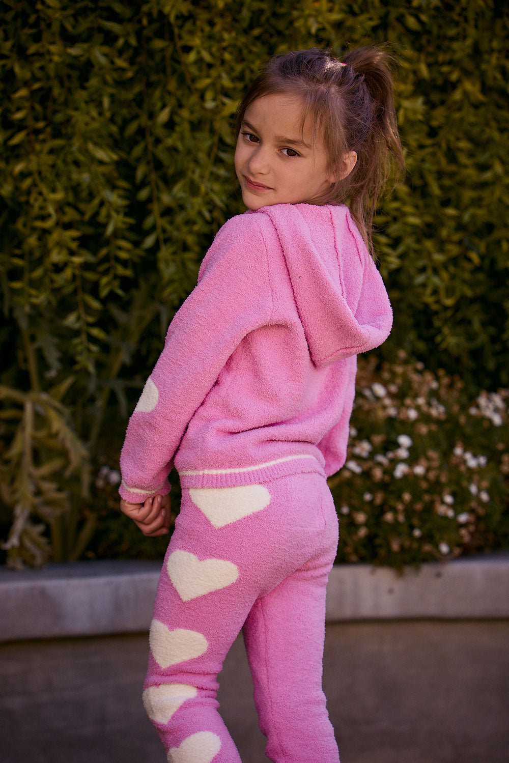 Chaser Kids Holiday Hug Girls Hoodie in pink, rear three-quarter view with hood down outdoors beside a low wall and greenery