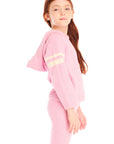 Side view of girl wearing Chaser's Cropped Shirred Confetti Pullover pink Hoodie and matching pants on a white background
