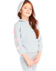 Girl wearing Chaser's light blue Cropped Shirred Snowflake Pullover Hoodie, hood up, with pink hearts on the sleeves on a white background