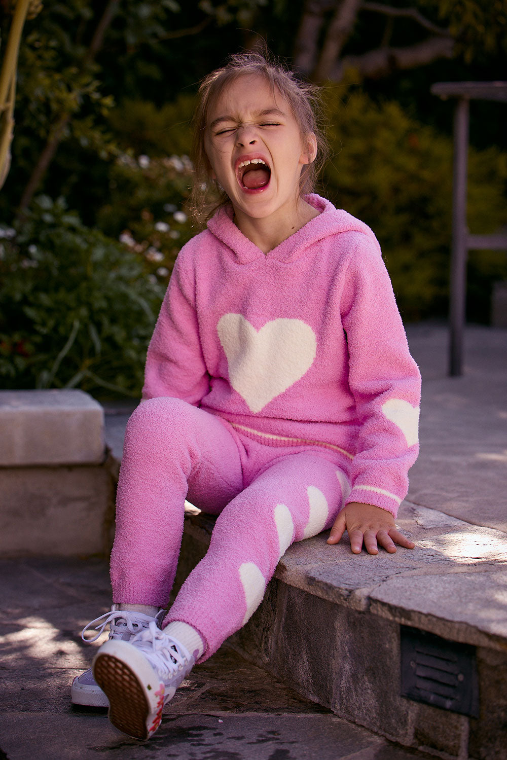 Chaser Kids Holiday Hug Girls Pants in pink with white hearts, worn by a girl seated on a low wall outdoors beside greenery